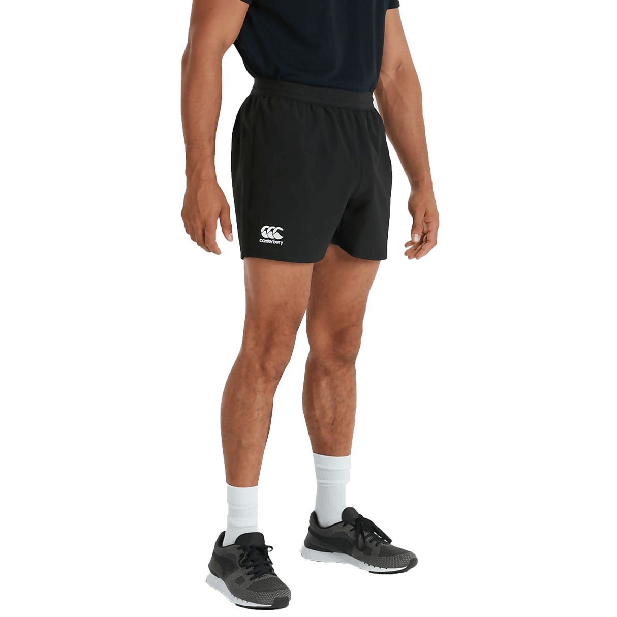 Man wearing a black t-shirt, Canterbury Tournament Shorts with a white logo, white socks, and black athletic shoes, standing against a white background.
