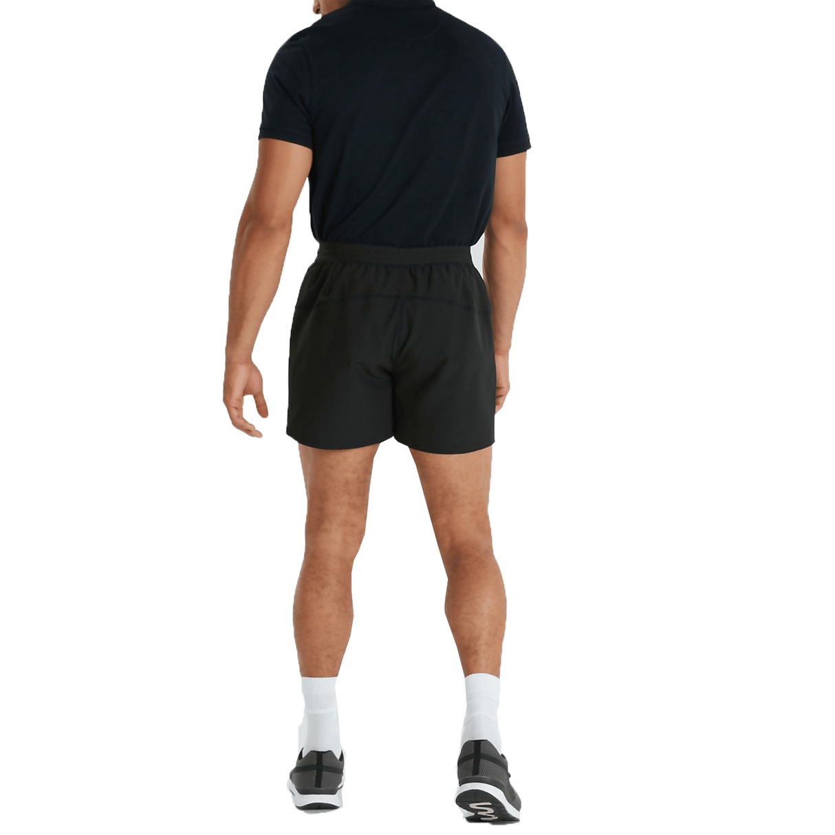 A man stands facing away, wearing a black shirt, Canterbury's Canterbury Tournament Shorts made of ripstop material, white socks, and black sneakers.