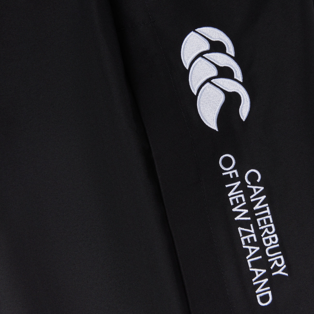 Close-up of black fabric with "Canterbury" branding and a white logo featuring three stylized shapes. The Women's Canterbury Open Hem Stadium Pant is perfect for rugby training, offering full-length leg zips for easy wear and versatility.