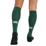 A person wearing Canterbury Playing Socks from Canterbury—green knee-high rugy socks featuring a white logo on the calf and black feet, along with a cushioned foot bed—is shown from behind.