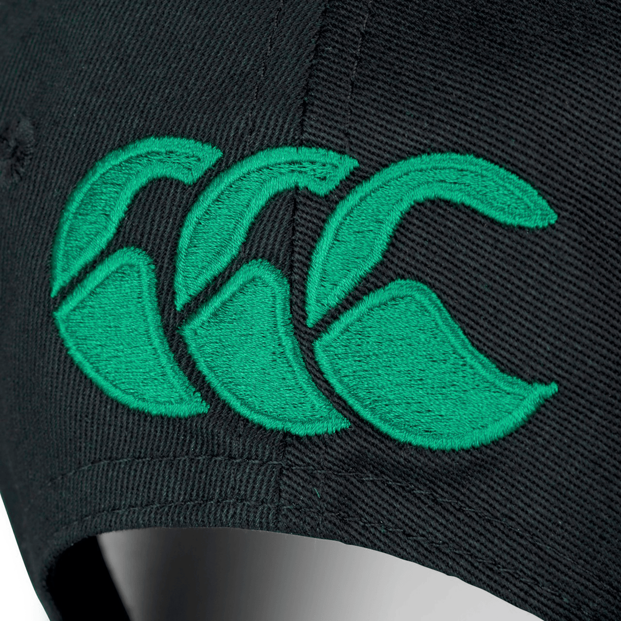 Close-up of a black fabric material with green Ireland logo embroidery, featuring three stylized shapes that resemble the letters "CCC" from the Ireland 24 Drill Cap by Canterbury.