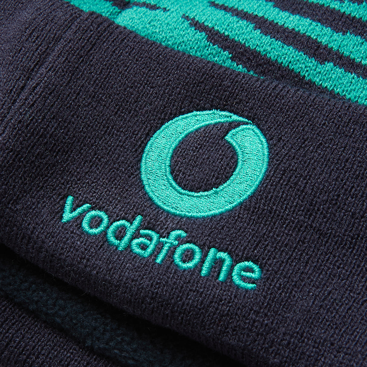 Close-up of a dark blue fabric with the Vodafone logo and text embroidered in teal, featured on the Ireland 26 Bobble Hat by Canterbury.