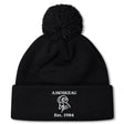 The Amoskeag Rugby Club Pom Pom Beanie by EMB Canterbury is a black knit hat with a folded brim, featuring “AMOSKEAG,” a ram logo, and “Est. 1984” embroidered in white.