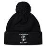 The Amoskeag Rugby Club Pom Pom Beanie by EMB Canterbury is a black knit hat with a folded brim, featuring “AMOSKEAG,” a ram logo, and “Est. 1984” embroidered in white.