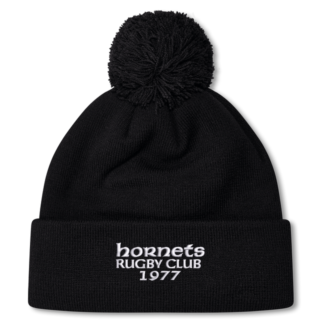 The Hornets Rugby Club 1977 Pom Pom Beanie by Canterbury from EMB Canterbury features a black knit design in the classic Canterbury bobble hat style, with a pom-pom on top and white embroidered "hornets RUGBY CLUB 1977" text on the front cuff.