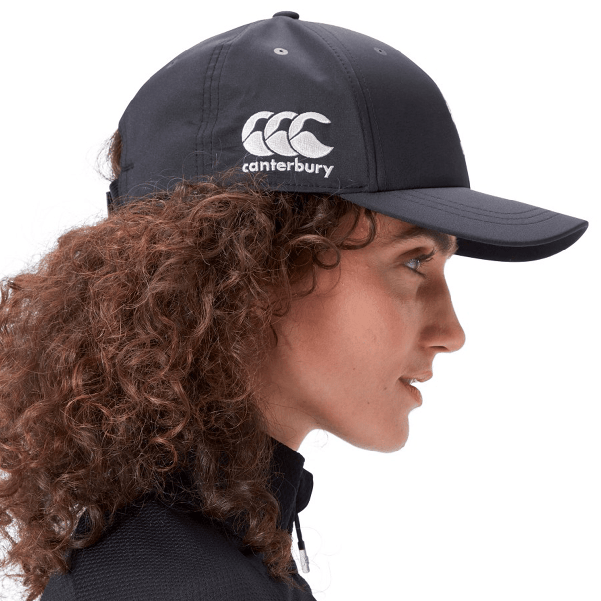 Side profile of a person wearing the Ireland 24 Tech Logo Cap by Canterbury, showcasing an adjustable strap and the Canterbury logo on the side. The person has curly brown hair and is dressed in a black jacket.