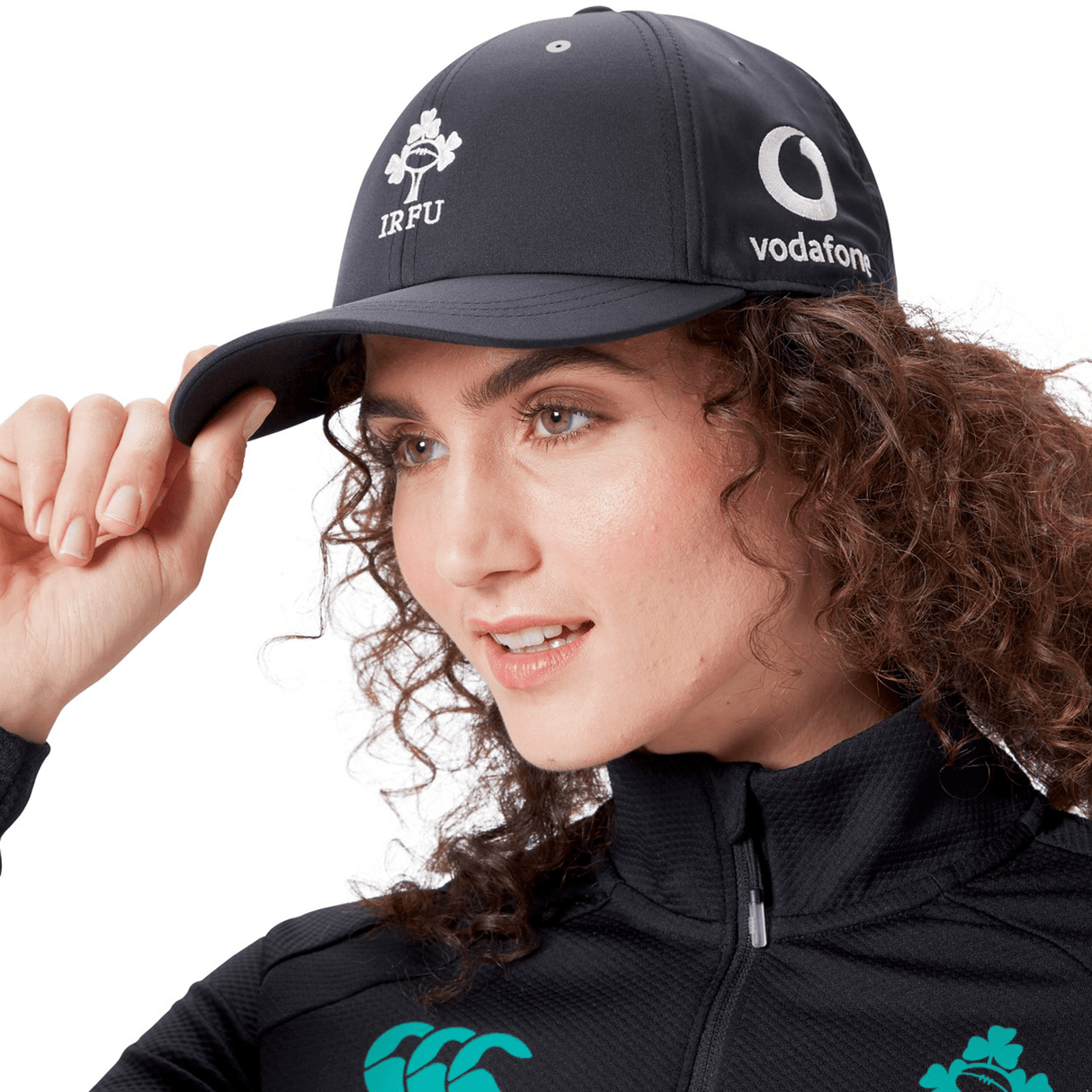A person wearing a black sports cap with the IRFU and Vodafone logos, featuring an adjustable strap, and a black sports jacket with teal logos, adjusts the brim of their Ireland 24 Tech Logo Cap by Canterbury.