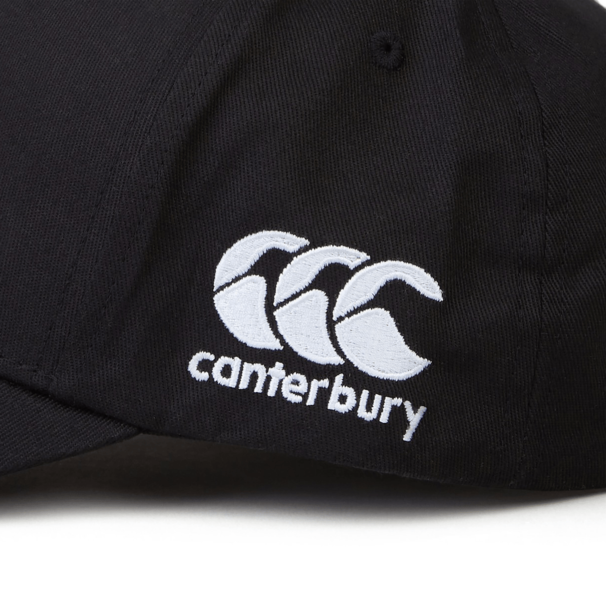 Close-up of a black Fiji Shield Flex Drill Cap by EMB Canterbury, showcasing an embroidered logo with three overlapping white curves and "Canterbury" underneath. Newly released, this cap combines classic style with modern flair, ideal for any wardrobe.