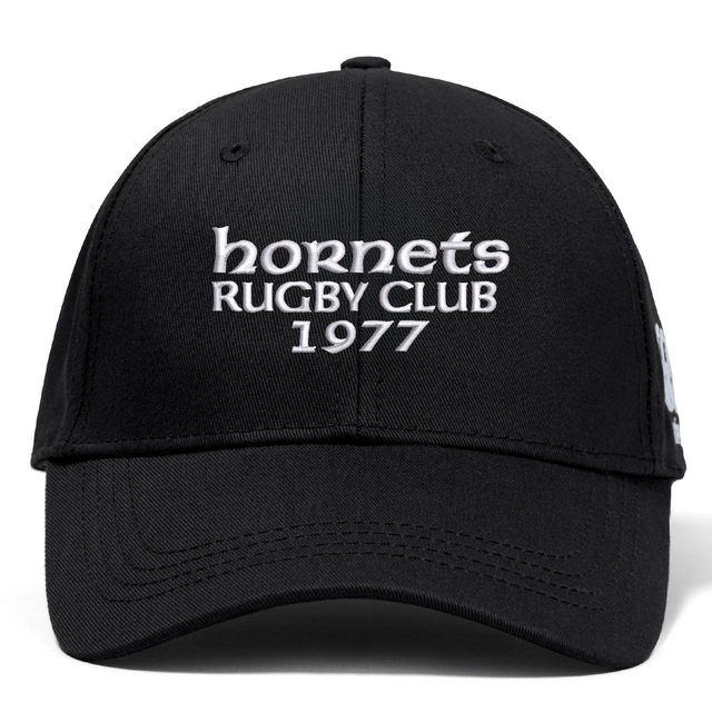The Hornets Rugby Club 1977 Flexfit Drill Cap by Canterbury features black fabric with white embroidered "hornets RUGBY CLUB 1977" text on the front. This curved brim cap from EMB Canterbury also includes a subtle CCC logo, combining style and comfort for rugby fans.