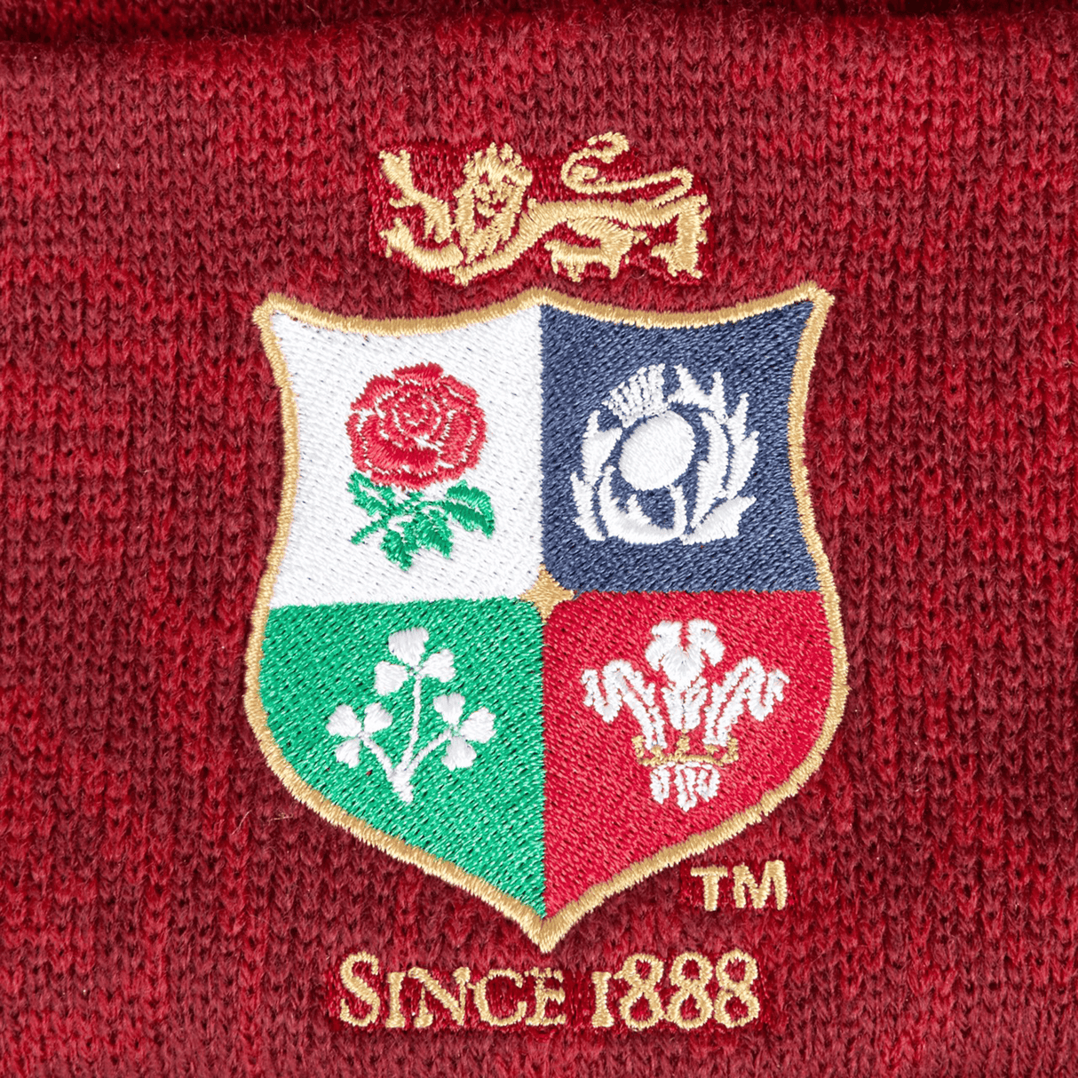 A close-up of the red dahlia-colored British & Irish Lions 2025 Pom-Pom Beanie by Canterbury showcases an embroidered lion logo above a shield decorated with four emblems: a rose, thistle, shamrock, and feathers. Beneath is the text "Since 1888," highlighting the longstanding heritage of the British & Irish Lions.