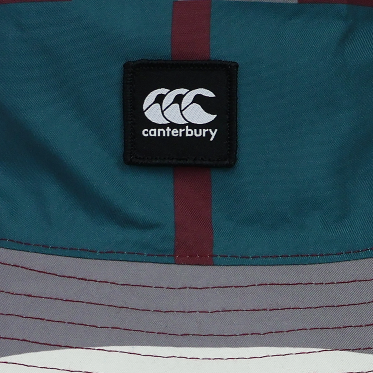 Close-up of a Canterbury logo patch on the Canterbury Uglies Red Reversible Bucket Hat, showcasing multicolored teal, maroon, grey, and white sections in a stylish reversible design.