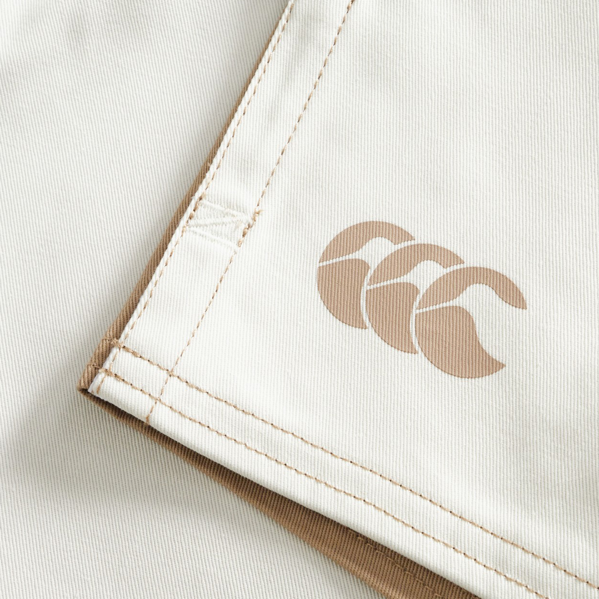 Close-up of Canterbury Fundamental Harlequin Drill 3" Shorts in light beige, featuring tan stitching and a tan three-wave logo printed on the surface.