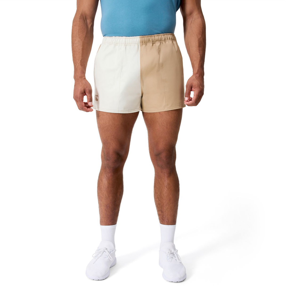 A person wearing a blue shirt, Canterbury Fundamental Harlequin Drill 3" Shorts in beige and off-white, white socks, and white sneakers stands against a white background.