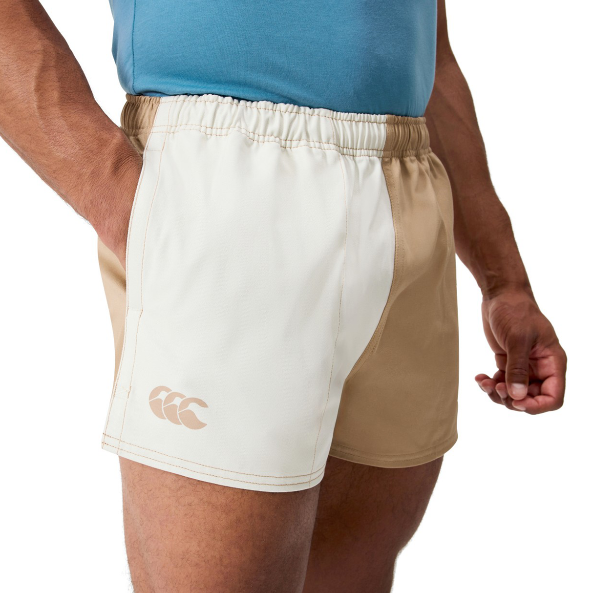 A person wearing Canterbury Fundamental Harlequin Drill 3" Shorts featuring a white front panel, beige side panels, and a visible Canterbury logo, standing with one hand by their side.