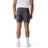 A person stands facing away on a plain white background, wearing a white shirt, white socks, white shoes, and dark gray Everest Woven Shorts by Canterbury.