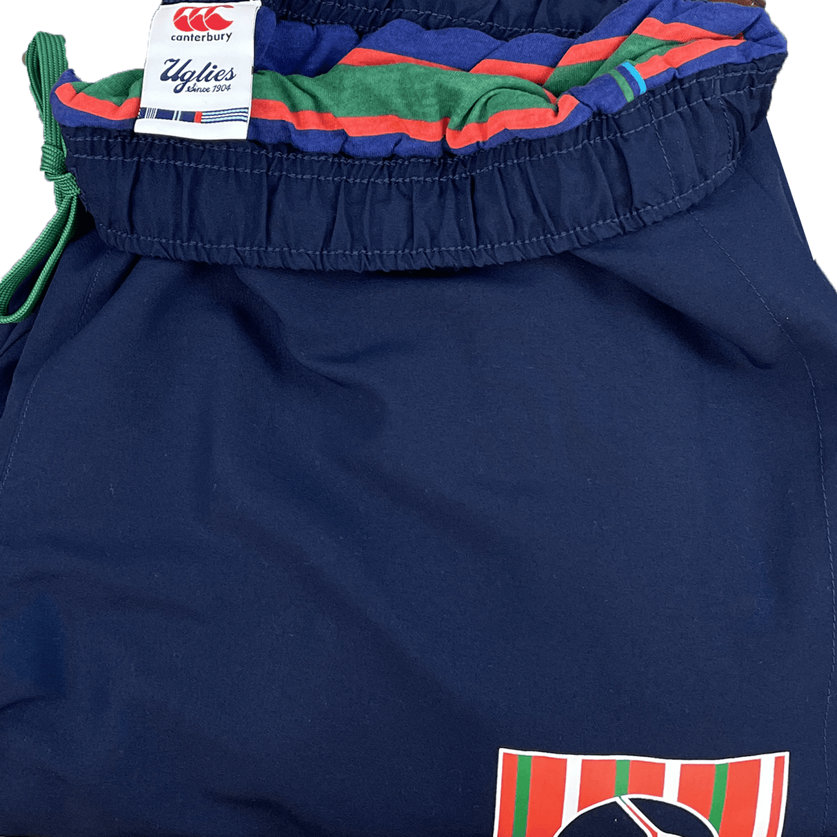 Introducing the Canterbury CCC Uglies Tapered Cuff Stadium Pant in Peacoat. These navy blue pants feature a stylish green, red, and white striped waistband. A small crest with matching stripes is visible on the lower part of the leg, showcasing distinctive Canterbury branding for added comfort.