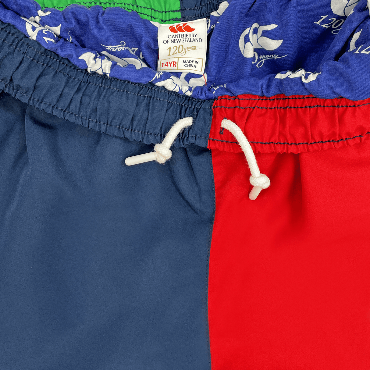 Close-up of a Canterbury Youth Harlequin Stadium Trackpant 24 with red, blue, and green panels featuring a white drawstring. The label reads "Canterbury," size "14YRS," echoing the vibrant design of the Harlequin Trackpant.