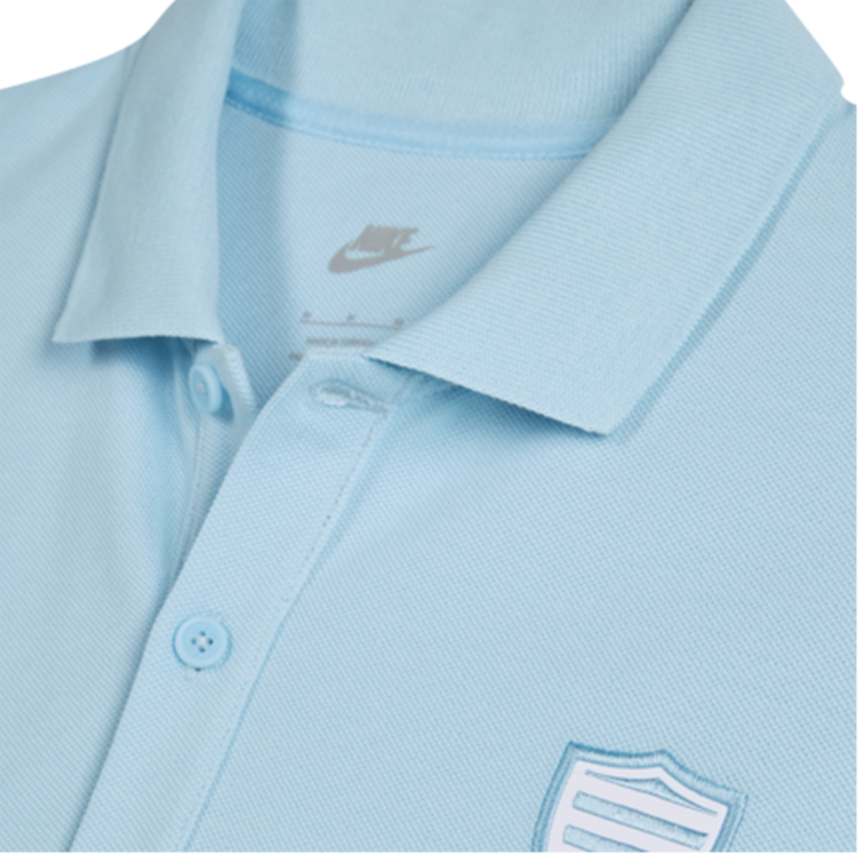 Close-up of the Nike Racing 92 25/26 Pique Polo in light blue, crafted from breathable cotton with buttons and a shield-shaped embroidered chest patch.