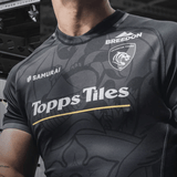 Close-up of a person wearing a black breathable jersey with "Leicester Tigers Change Jersey 23/24 by Samurai" and other logos, standing in a gym.