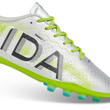 The Ida Women's Rise Club FG "Play All Day" by IDA Sports is a silver and green firm ground cleat with lime green laces and studs, made from vegan leather, featuring "IDA" on the side.