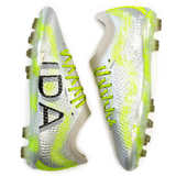 A pair of Ida Women's Rise Elite FG "Play All Day" soccer cleats by IDA Sports in silver and green featuring textured surfaces for enhanced ball control, with black lettering on the side and neon yellow laces.