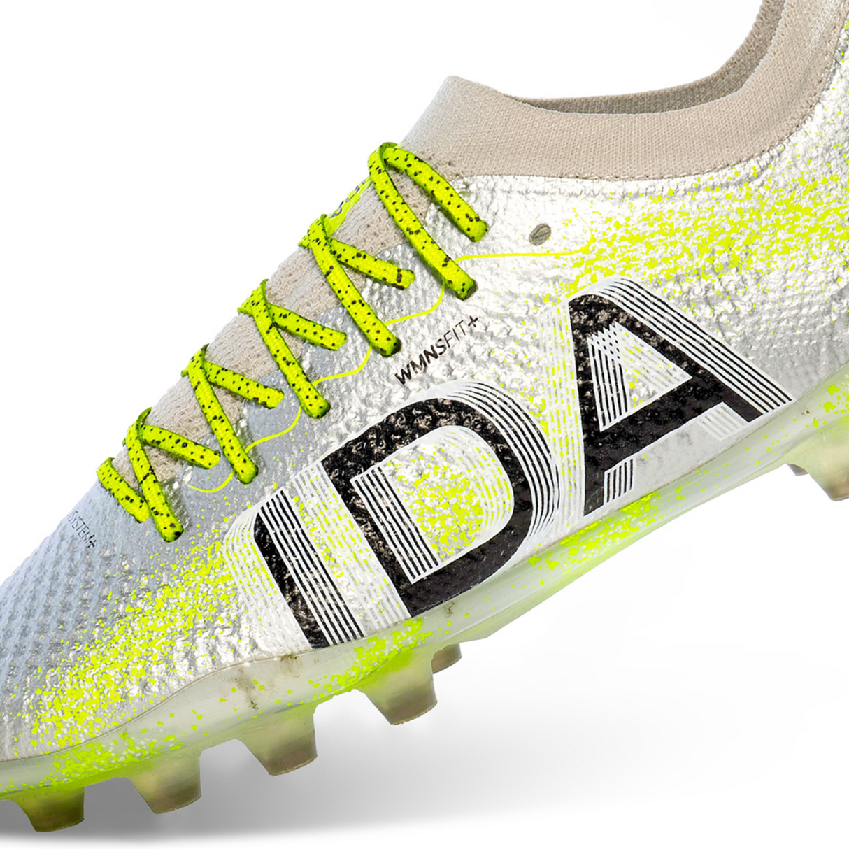 Close-up of the Ida Women's Rise Elite FG "Play All Day" cleat in silver/green by IDA Sports, showcasing WAVEKNIT for flexibility, textured surfaces with "IDA," and bright green laces and spikes for traction. Made from 100% vegan materials.