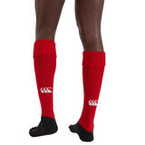 Person wearing Canterbury Playing Sock knee-high rugby socks in bright red, featuring black soles, a white Canterbury logo on the back, and a cushioned foot bed, viewed from behind.