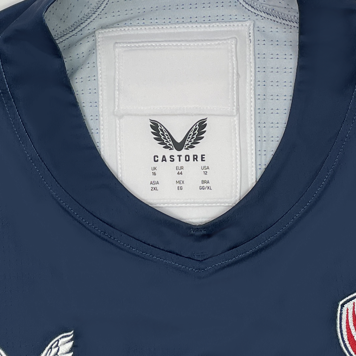 Close-up of a Women's USA Rugby Pro Home Jersey by Castore shows a navy blue label with international sizes and highlights its moisture-wicking fabric.