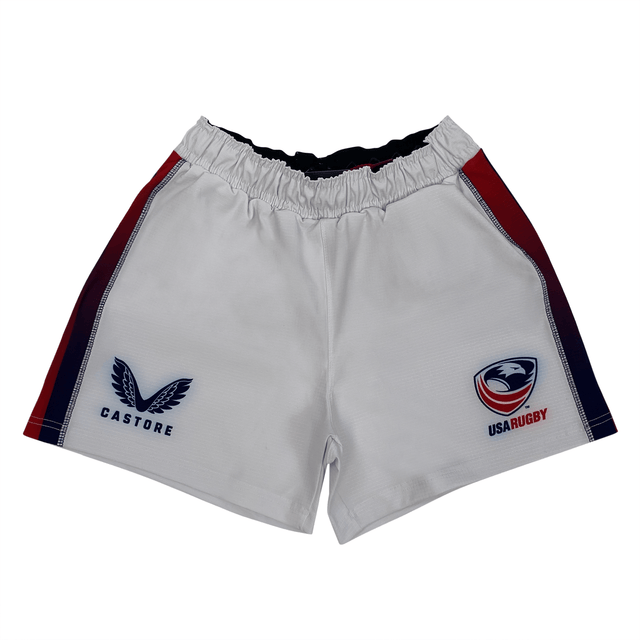 Women's USA Rugby Pro Home Shorts by Castore feature a moisture-wicking fabric, an elasticated waistband, and are adorned with the "CASTORE" logo on the left and "USA RUGBY" emblem on the right. Stylish red and blue stripes run down the sides.