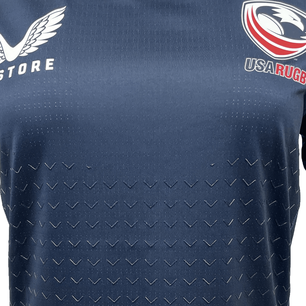 The Women's USA Rugby Training Jersey by Castore, in navy blue, features an embossed chevron pattern with winged and eagle logos. Made from moisture-wicking fabric, it displays "STORE" and "USA RUGBY" on the chest and offers a Women's Athletic Fit for comfort and style.