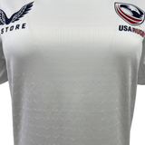 Close-up of a Castore Women's USA Rugby Training Jersey, showcasing blue USA Rugby and eagle graphics on a white athletic fit. It features moisture-wicking fabric for optimal performance.