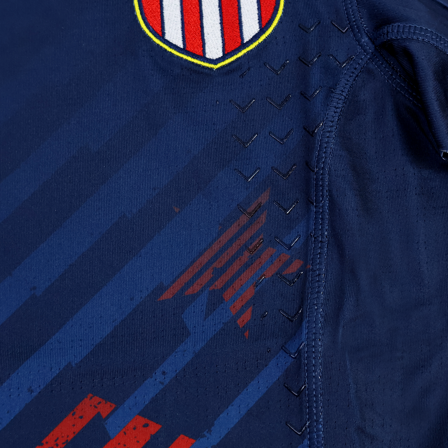Close-up of the Women's USA Rugby 24/25 7s Olympic Pro Home Jersey by Castore, in navy blue with a shield emblem featuring red and white stripes, plus chevron-shaped ventilation holes along the seam.