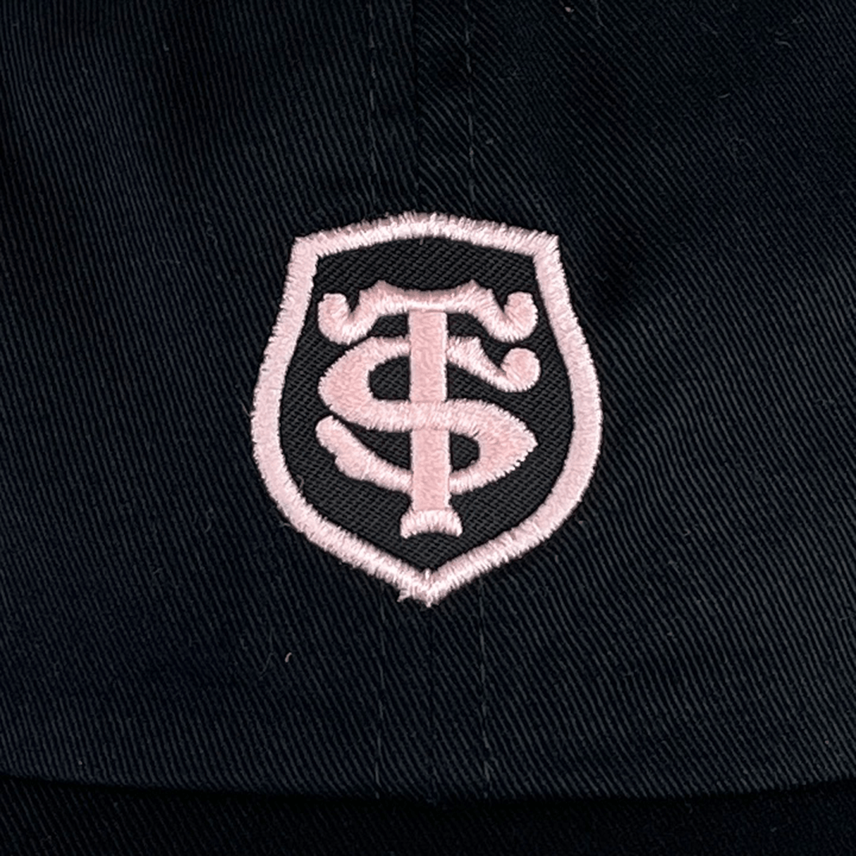 A black and pink adjustable hat features a white embroidered shield-like emblem with stylized letters "TS" intertwined, reminiscent of the Stade Toulousain Heritage 86 Cap by Nike design.