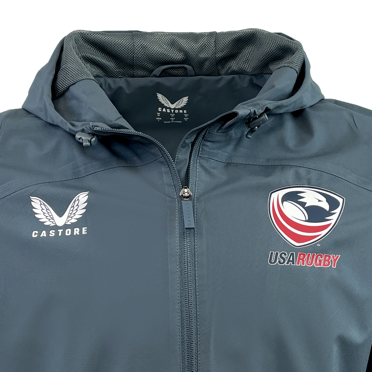The USA Rugby Rain Jacket by Castore in dark gray features a hood, Castore logo on the left, USA Rugby emblem on the right, and is crafted with water-resistant technology and a lightweight mesh lining for comfort in unpredictable weather.