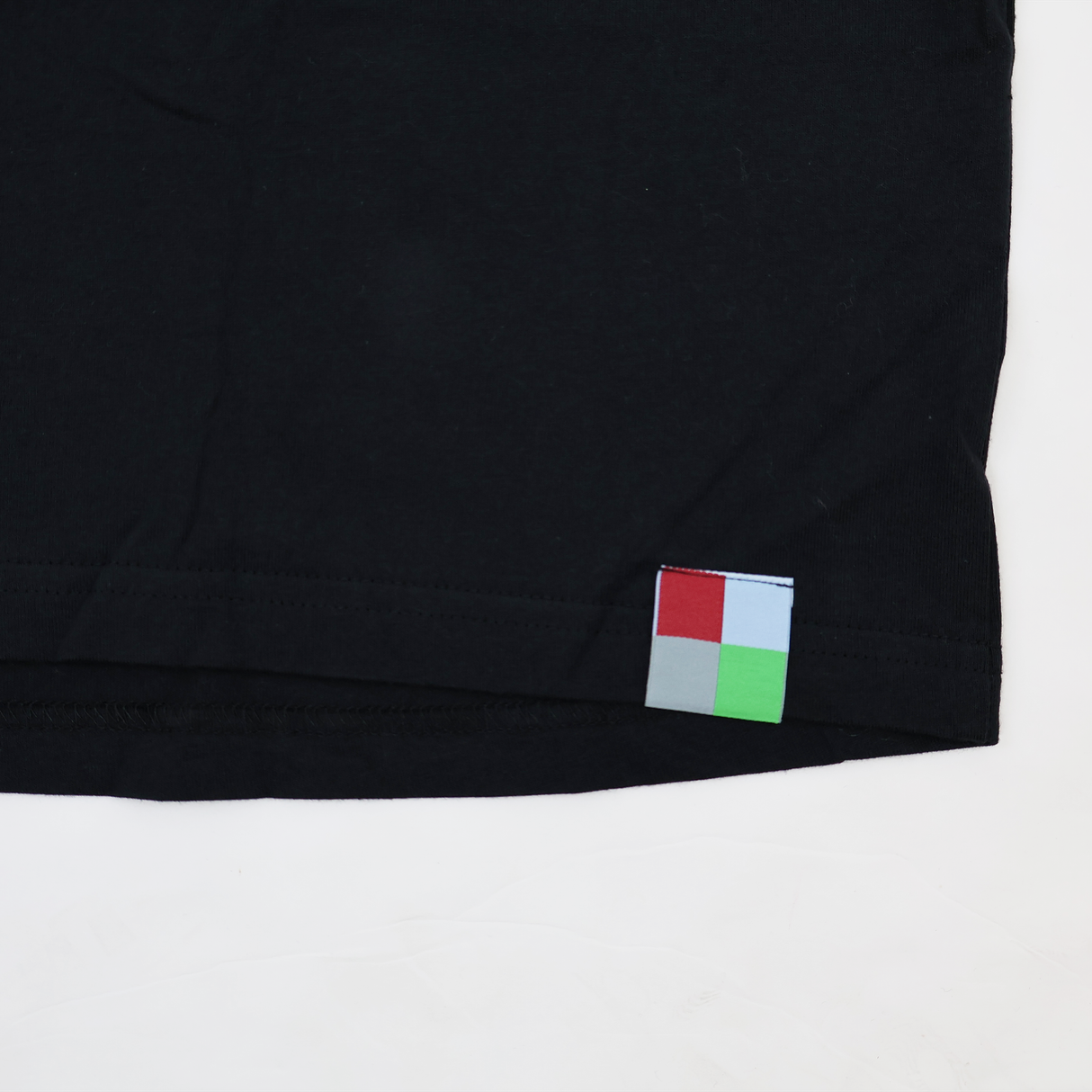 A close-up of the hem of a black Castore Harlequins 22/23 Heritage Cotton Tee, showing a small square tag with red, light blue, gray, and green quadrants.