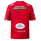 The Scarlets Replica Home Jersey 23/24 by Castore is a red athletic fit jersey adorned with various sponsor logos on the back, including Ascona, Dyfed Steels, Pencnwc Holiday Park, and Colegsirgar. This Welsh rugby union jersey is perfect for fans.