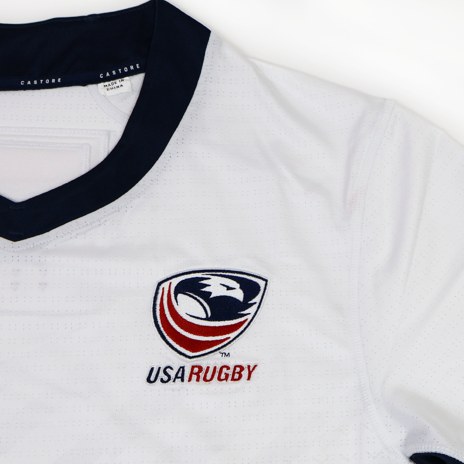 The USA Rugby 24/25 Pro Away Jersey by Castore features a white design with a dark collar, bold red and navy lettering on the chest, and the USA Rugby eagle logo.