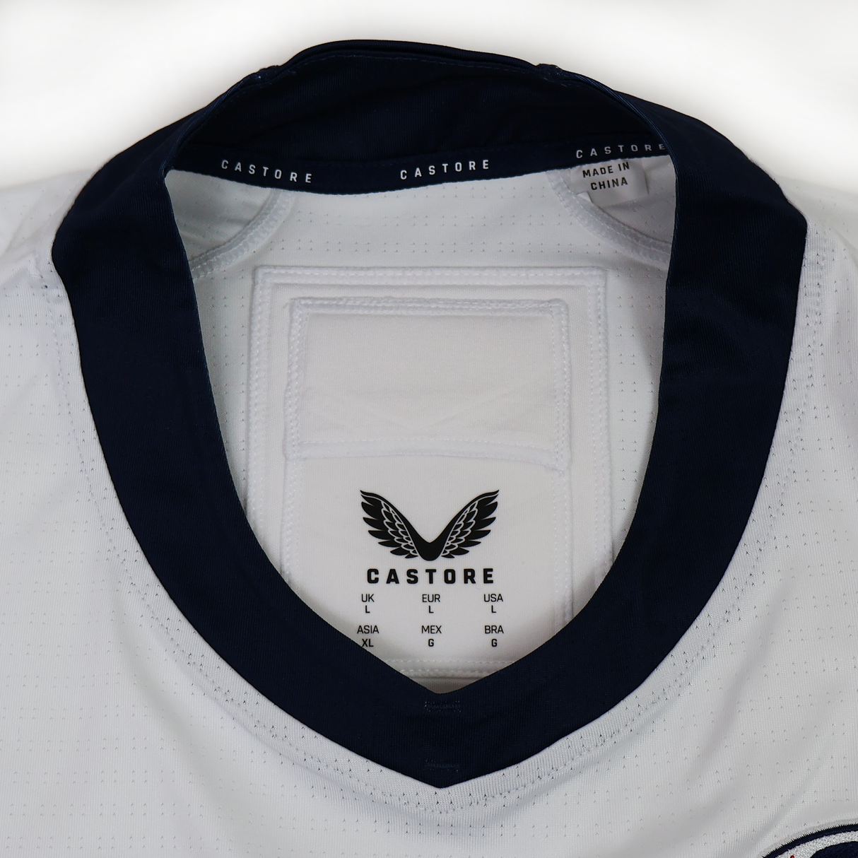 The USA Rugby 24/25 Pro Away Jersey by Castore features white fabric with a navy collar, showcasing the Castore logo, size chart, and a "Made in China" label inside the neck.