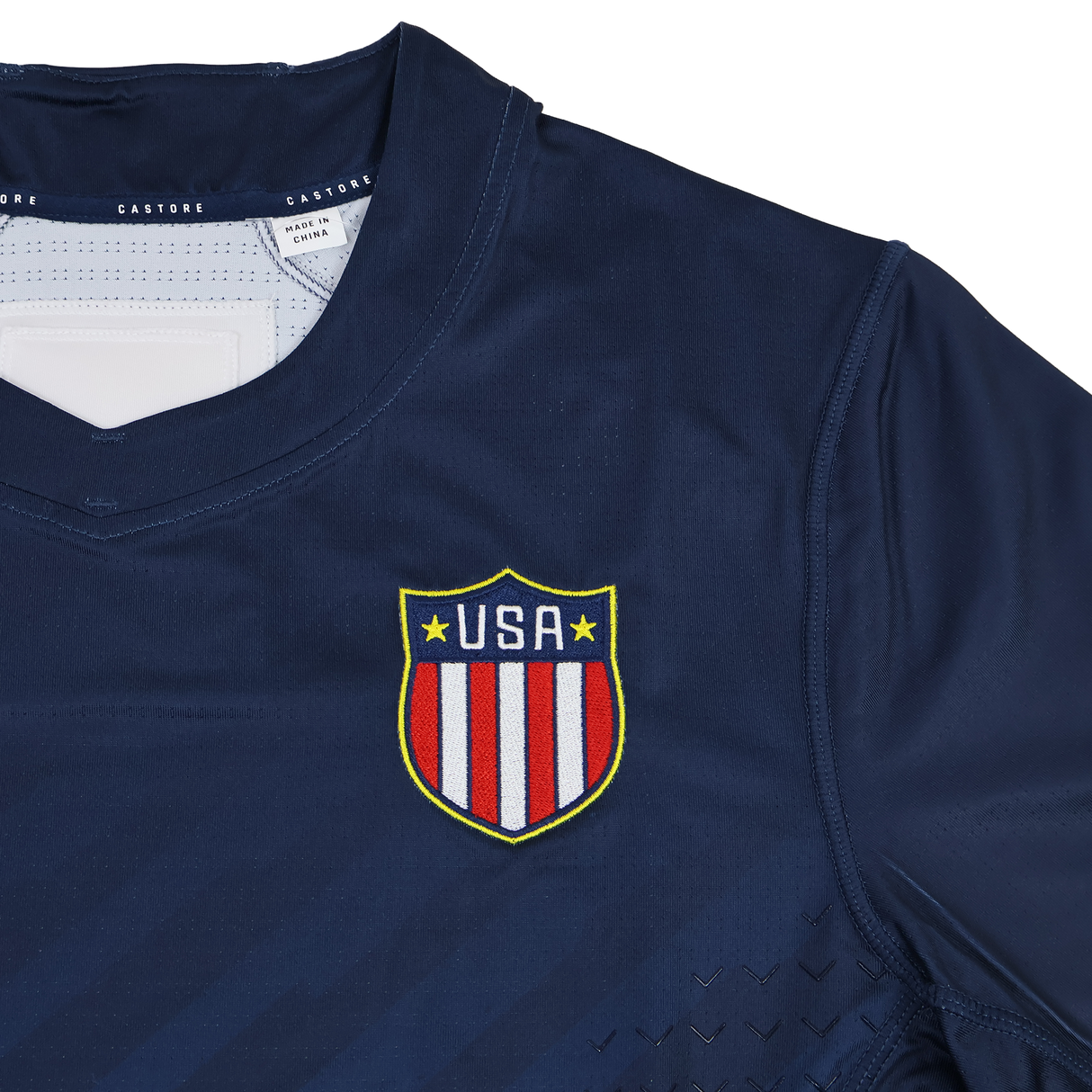 The USA Rugby 24/25 7s Olympic Pro Home Jersey by Castore features a navy blue design with a shield emblem of red and white stripes and yellow stars on the chest, inspired by Olympic style.