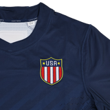 The USA Rugby 24/25 7s Olympic Pro Home Jersey by Castore features a navy blue design with a shield emblem of red and white stripes and yellow stars on the chest, inspired by Olympic style.
