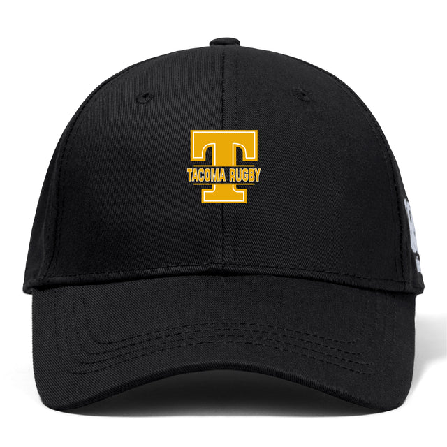 The EMB Canterbury Tacoma Tsunami Rugby Flexfit Drill Cap features a black design with a gold "T" and "TACOMA RUGBY" embroidered on the front.