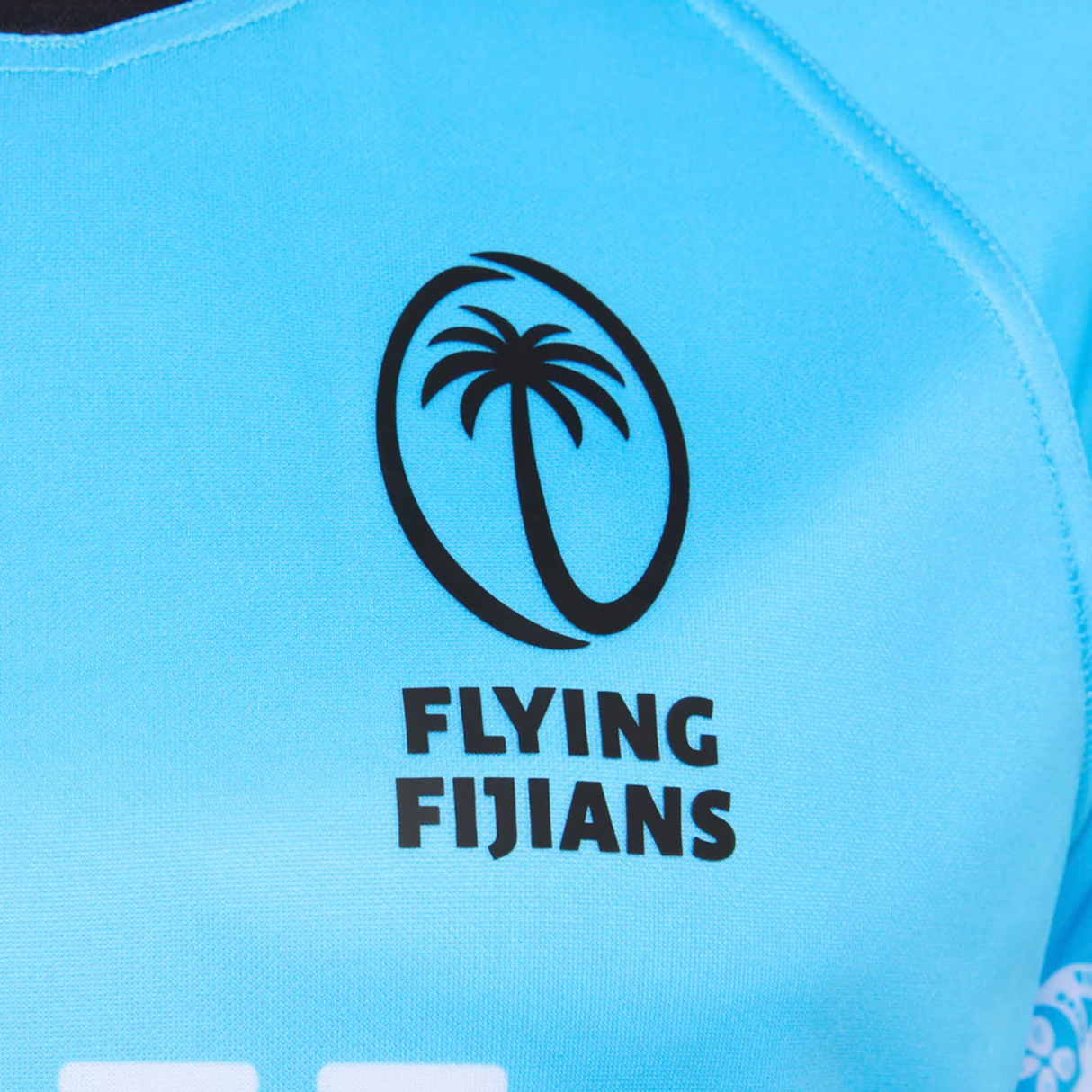 Close-up of the Fiji 25/26 Replica Away Jersey by Umbro, a light blue polyester athletic fit featuring the Flying Fijians logo, stylized palm tree, bold black text, and intricate Fijian masi art details.