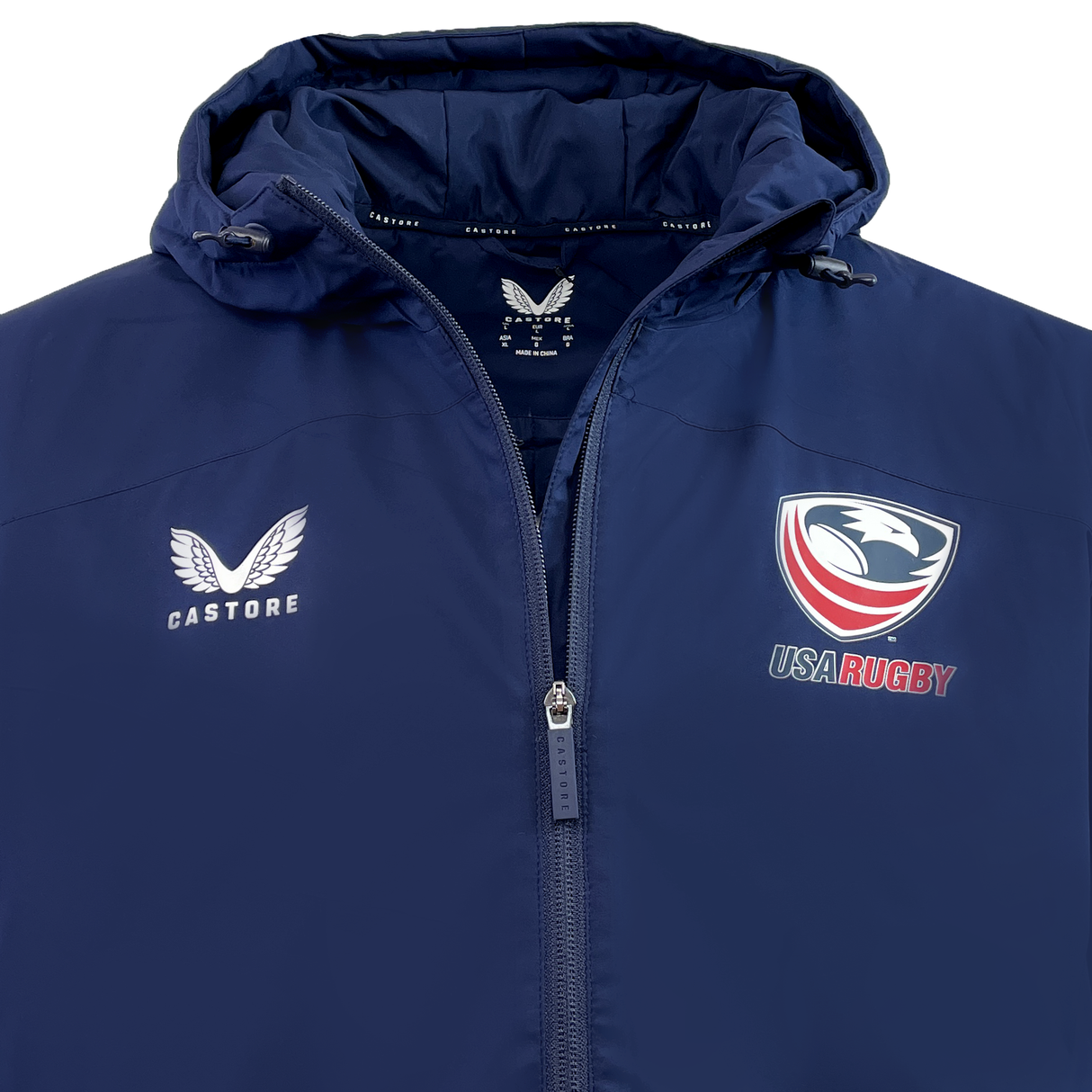 The USA Rugby Long Length Bench Jacket by Castore is a navy blue jacket featuring the Castore logo on the left side and the USA Rugby logo on the right. It includes a front zipper and a hood, making it ideal for staying warm while showing team support.