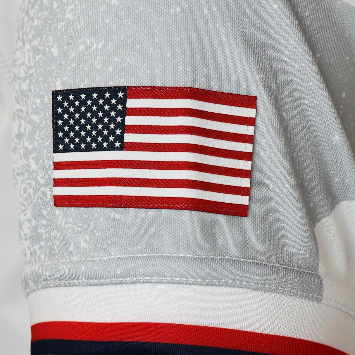 A close-up of a gray fabric sleeve with an embroidered United States flag patch, part of the Women's USA Rugby WRWC25 Replica Away Jersey by Macron.
