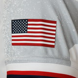 A close-up of a gray fabric sleeve with an embroidered United States flag patch, part of the Women's USA Rugby WRWC25 Replica Away Jersey by Macron.