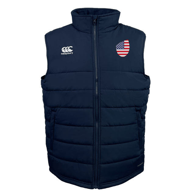 The USA Shield Core Gilet by EMB Canterbury is a navy blue puffer vest with an American flag patch on the left chest, a company logo on the right chest, Thermoreg insulation, and a front zipper.