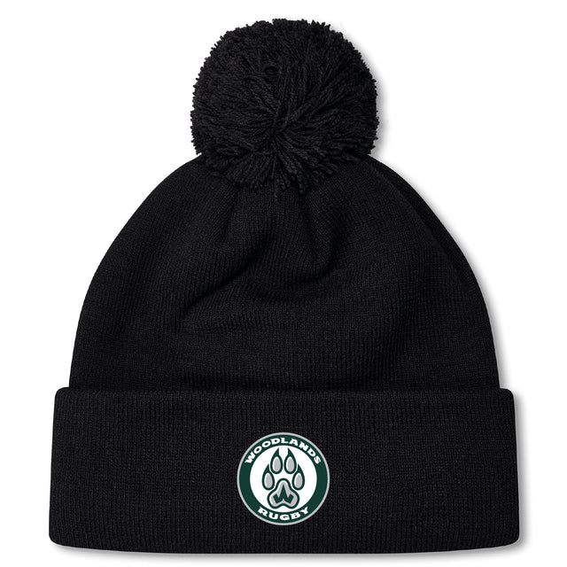The Woodlands Youth Pom Pom Beanie by EMB Canterbury is a black, fleece-lined hat with a pom-pom and a "Woodlands Rugby" paw print logo patch—perfect for keeping warm at chilly rugby games.