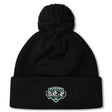 The Woodlands Youth Wolfpack Pom Pom Beanie by Canterbury from EMB Canterbury is a black knit, fleece-lined beanie with a large pom-pom and a "Woodlands Rugby" logo patch with three wolves on the folded brim.