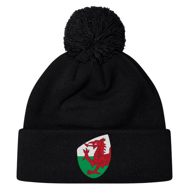 The EMB Canterbury Wales Shield Pom Pom Beanie is a stylish black beanie with a striking patch featuring a red dragon against a green and white backdrop, reminiscent of the Wales Shield.