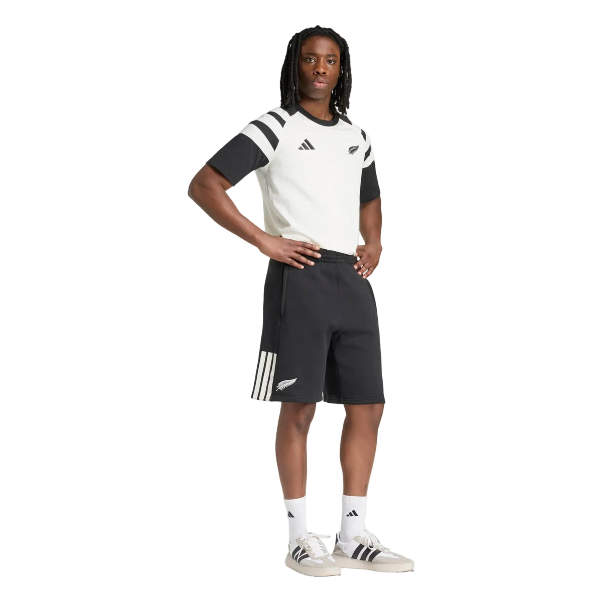 A person stands with one hand on their hip, dressed in black and white adidas sportswear featuring the All Blacks 26/27 Leisurewear Shorts by adidas, paired with matching adidas shoes and socks, against a plain white background.